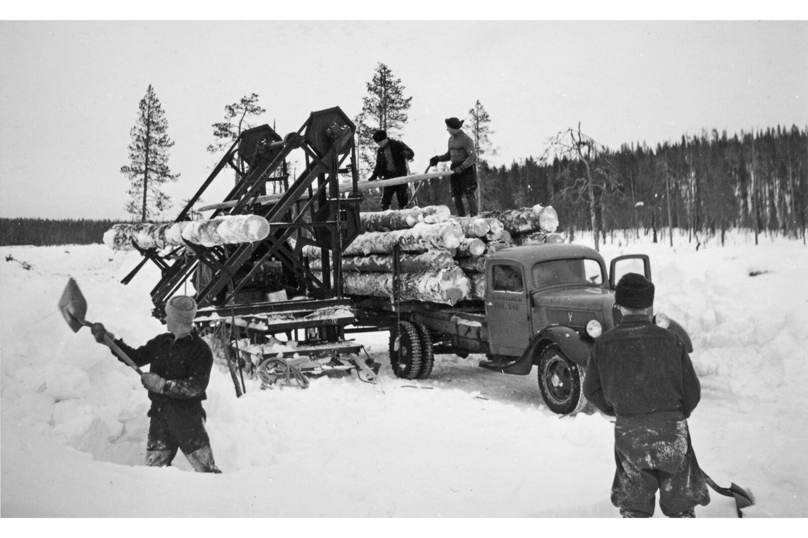 Kuormurin talvirenkaat tositoimissa lumisessa maastossa. Henkilöitä kaivamassa lunta pois kuormurin tieltä.