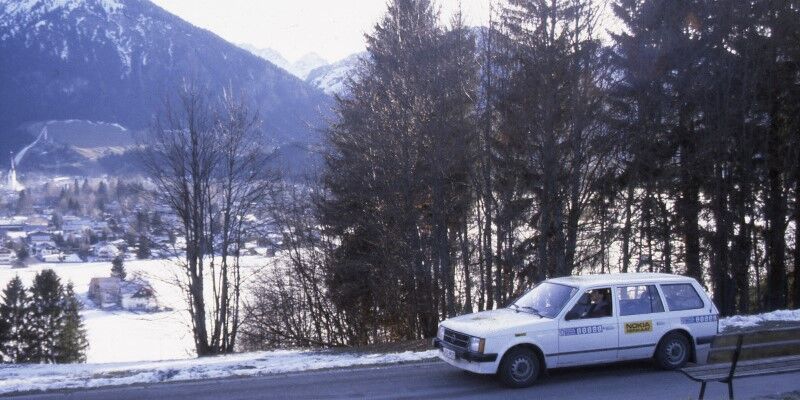 A white car driving in front of a row of trees, mountains in the background